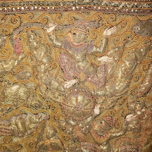 May include: Close-up of an ornate textile with gold and bronze embroidery. The design features figures in traditional garb, with intricate detailing and embellishments. The textile has a rich, textured appearance, with a variety of patterns and colors.