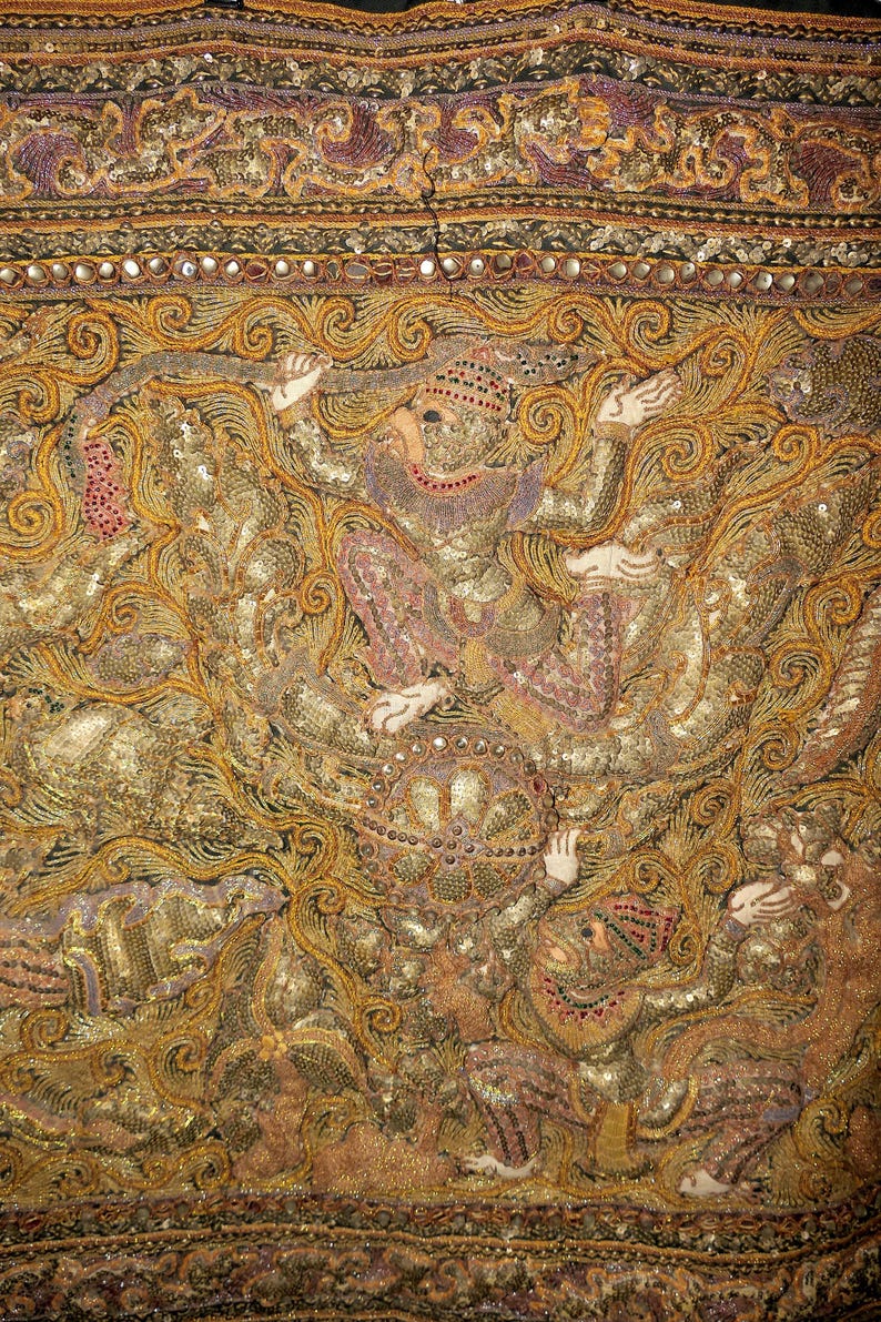 May include: Close-up of an ornate textile with gold and bronze embroidery. The design features figures in traditional garb, with intricate detailing and embellishments. The textile has a rich, textured appearance, with a variety of patterns and colors.