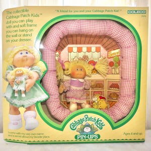 May include: A Cabbage Patch Kids Pin-Ups collectible doll in original packaging. The doll has blonde yarn hair in pigtails, a purple and white checkered dress, and white shoes. It is framed by a pink and white gingham padded border.
