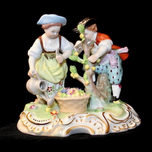 Puede incluir: Un conjunto de figuras de porcelana que representa a dos figuras, una mujer con una regadera y un hombre, cosechando fruta de un &aacute;rbol. Las figuras est&aacute;n vestidas con ropa colorida y se encuentran sobre una base verde con detalles dorados.