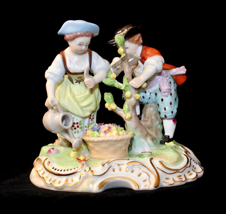 Puede incluir: Un conjunto de figuras de porcelana que representa a dos figuras, una mujer con una regadera y un hombre, cosechando fruta de un &aacute;rbol. Las figuras est&aacute;n vestidas con ropa colorida y se encuentran sobre una base verde con detalles dorados.
