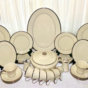 May include: A complete cream-colored dinnerware set with a silver trim. The set includes plates of various sizes, oval platters, cups, saucers, a covered serving dish, and salt and pepper shakers. The dishes are arranged on a white surface.