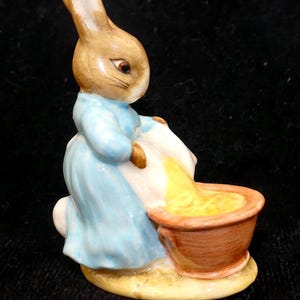 May include: A ceramic figurine of a brown rabbit wearing a light blue jacket and white apron, standing beside a small terracotta-colored bowl filled with a yellow substance, all set upon a light yellow oval base.