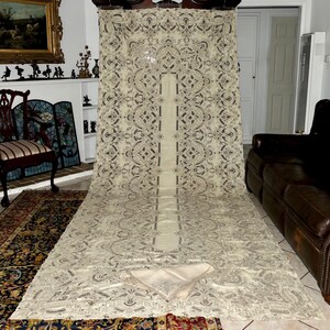 May include: A large, antique, white lace tablecloth with a delicate floral pattern. The tablecloth is laid out on a patterned rug and has a matching set of napkins.