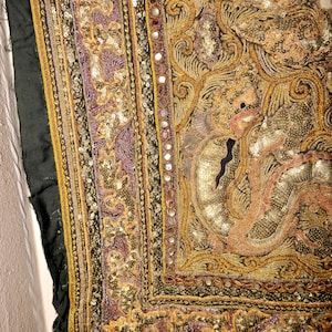 May include: Close-up of an ornate textile with intricate embroidery and embellishments. The design features a central figure with detailed patterns in gold, purple, and peach hues. The edges are framed with a black border and small mirrors.