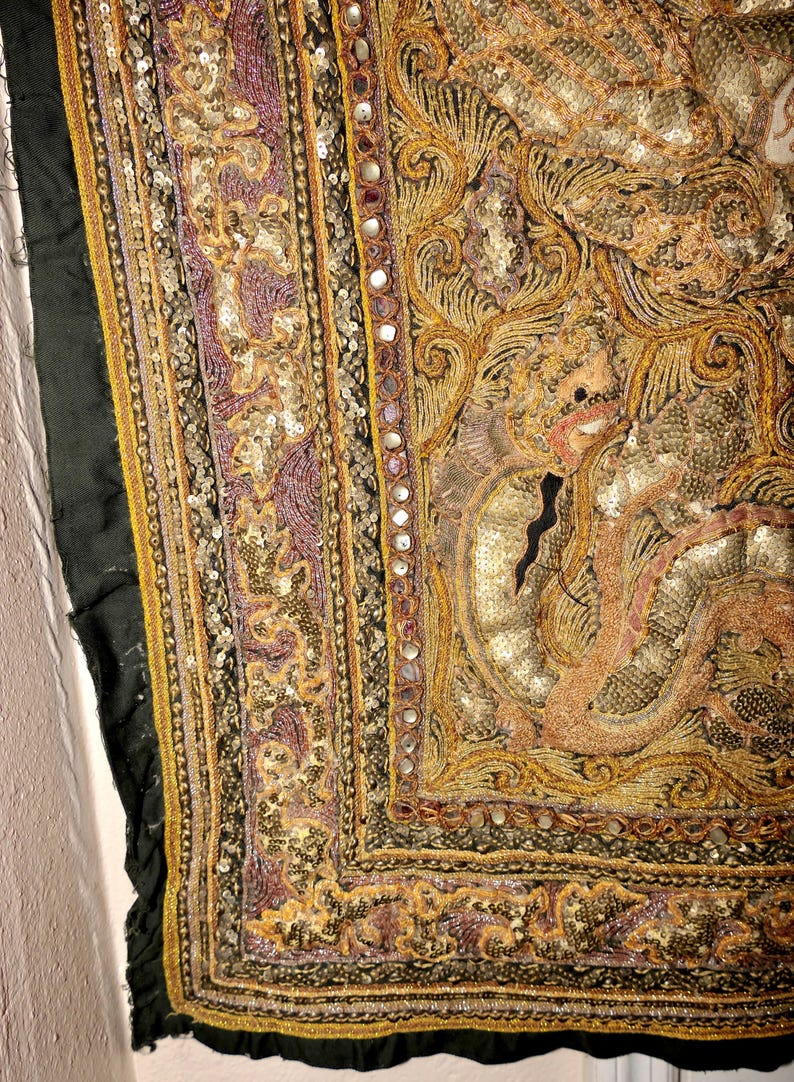 May include: Close-up of an ornate textile with intricate embroidery and embellishments. The design features a central figure with detailed patterns in gold, purple, and peach hues. The edges are framed with a black border and small mirrors.