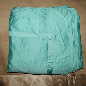 May include: A folded, solid light teal blanket with a smooth, slightly reflective finish. The blanket features a wide, stitched border detail along the edges, creating a subtle frame effect on the fabric surface. The material appears soft and lightweight.