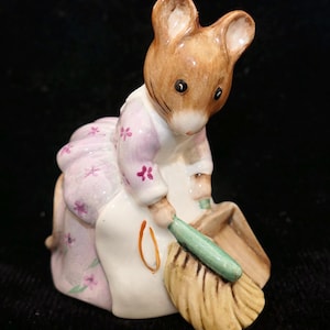 May include: A ceramic mouse figurine with brown fur, wearing a light pink dress with a small purple floral pattern and a white apron. The mouse is holding a green-handled broom and a tan dustpan, appearing to be in the act of sweeping.