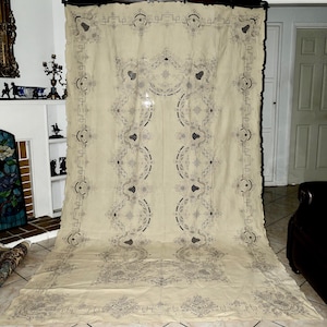 May include: A large, cream-colored vintage tablecloth with intricate gray floral and geometric embroidery. The edges are scalloped, and the design features repeating patterns and decorative borders. The tablecloth is displayed in a well-lit room.