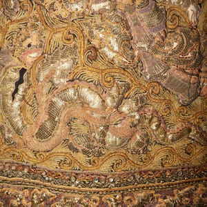 May include: Close-up of an ornate textile with intricate embroidery and embellishments. The design features gold, bronze, and pink tones, with detailed patterns and sequin accents. The textile has a black border.