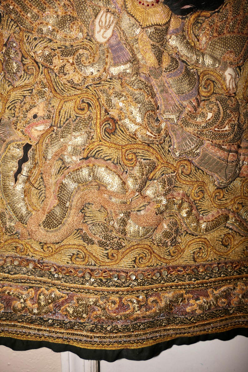 May include: Close-up of an ornate textile with intricate embroidery and embellishments. The design features gold, bronze, and pink tones, with detailed patterns and sequin accents. The textile has a black border.