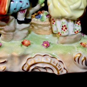 Puede incluir: Una figurita de porcelana de dos ni&ntilde;os, uno con un vestido rojo y azul y el otro con un vestido amarillo, de pie sobre una base verde con flores. La base tiene un borde dorado y una inscripci&oacute;n dorada.