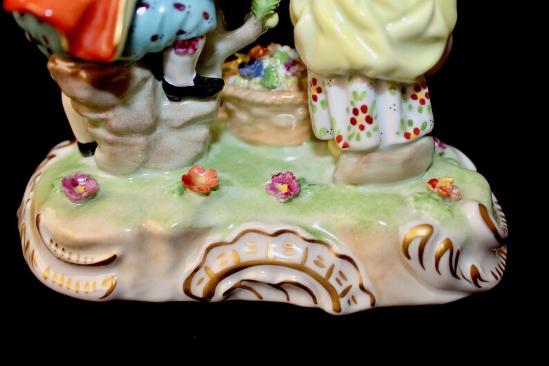 Puede incluir: Una figurita de porcelana de dos ni&ntilde;os, uno con un vestido rojo y azul y el otro con un vestido amarillo, de pie sobre una base verde con flores. La base tiene un borde dorado y una inscripci&oacute;n dorada.