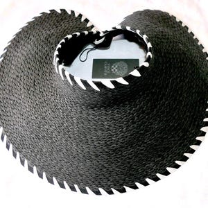 May include: A black straw visor with a wide brim featuring a white stitched edge detail. The inner band displays a white stitched trim and a black tag with the white text Vince Camuto printed on it. The visor has a woven texture throughout.