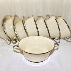 May include: A set of cream-colored soup bowls with silver trim and handles. The bowls are arranged in a neat stack, with one bowl in front. The bowls have a classic, elegant design, suitable for formal dining.