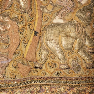 May include: Close-up of an ornate textile featuring an elephant and other figures, embellished with gold and bronze thread, sequins, and beads. The design includes intricate swirling patterns and a black border.