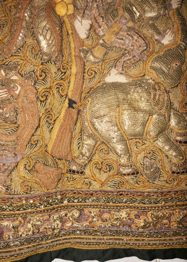 May include: Close-up of an ornate textile featuring an elephant and other figures, embellished with gold and bronze thread, sequins, and beads. The design includes intricate swirling patterns and a black border.