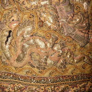 May include: Close-up of an ornate textile with intricate gold and bronze embroidery. The design features swirling patterns, sequins, and detailed figures, with a border of decorative trim. The fabric has a rich, textured appearance.