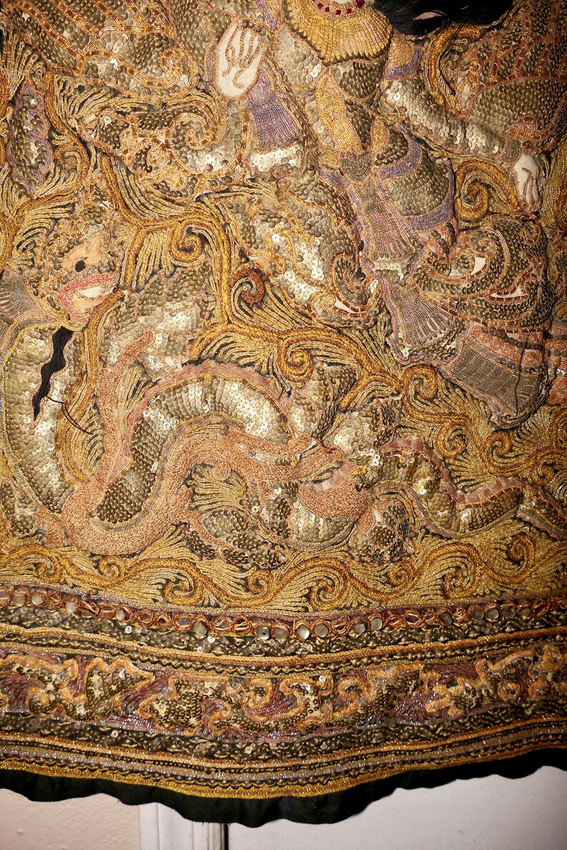 May include: Close-up of an ornate textile with intricate gold and bronze embroidery. The design features swirling patterns, sequins, and detailed figures, with a border of decorative trim. The fabric has a rich, textured appearance.