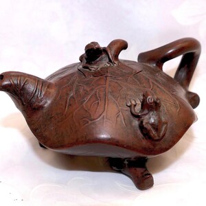May include: A brown, handcrafted teapot with a unique frog and leaf design. The teapot features a curved handle and spout, with a frog figurine on the lid and another on the side. The teapot rests on three small feet.