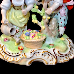 Puede incluir: Una figurita de porcelana de dos ni&ntilde;os cuidando un peque&ntilde;o &aacute;rbol con una cesta de flores en su base. Los ni&ntilde;os est&aacute;n vestidos con ropa colorida y la base de la figurita est&aacute; decorada con detalles dorados.