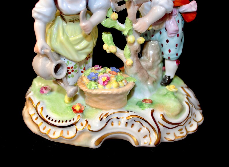 Puede incluir: Una figurita de porcelana de dos ni&ntilde;os cuidando un peque&ntilde;o &aacute;rbol con una cesta de flores en su base. Los ni&ntilde;os est&aacute;n vestidos con ropa colorida y la base de la figurita est&aacute; decorada con detalles dorados.