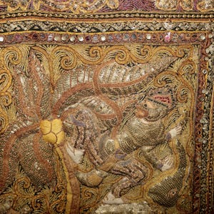 May include: Close-up of an ornate textile with intricate embroidery in gold, brown, and purple hues. The design features a detailed figure, possibly a deity or royal personage, surrounded by swirling patterns and embellishments.