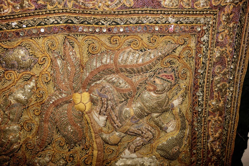 May include: Close-up of an ornate textile with intricate embroidery in gold, brown, and purple hues. The design features a detailed figure, possibly a deity or royal personage, surrounded by swirling patterns and embellishments.
