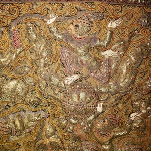 May include: Close-up of an ornate textile with gold and bronze embroidery. The design features figures in elaborate costumes, with intricate details and embellishments. The textile has a rich, textured appearance.