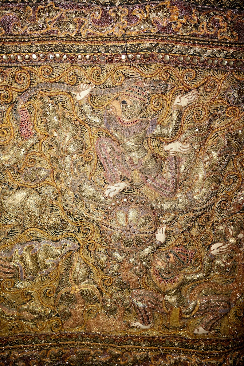 May include: Close-up of an ornate textile with gold and bronze embroidery. The design features figures in elaborate costumes, with intricate details and embellishments. The textile has a rich, textured appearance.