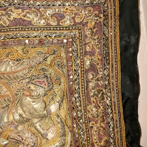 May include: An ornate textile with intricate embroidery and embellishments. The design features gold, purple, and silver threads, with small mirrored accents. The artwork depicts a figure in a detailed, textured style, set against a dark green background.