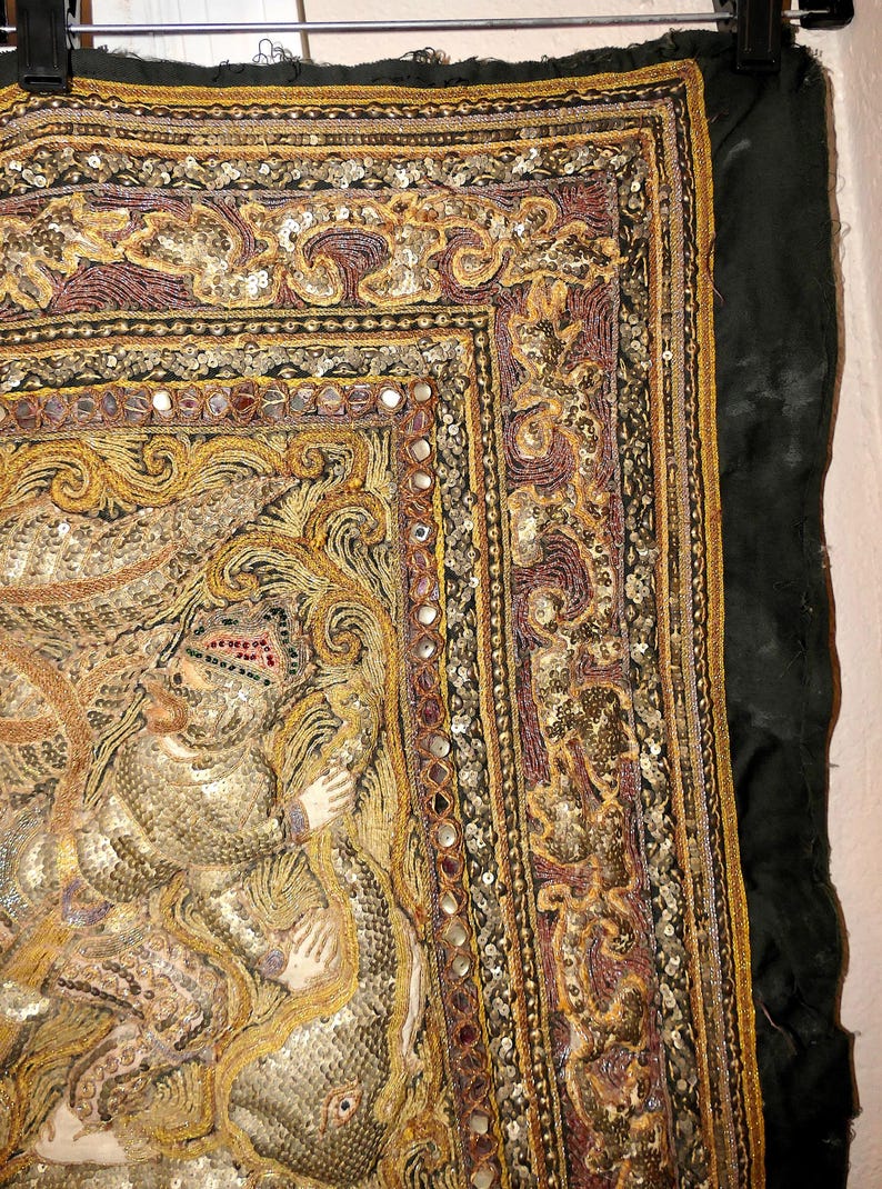 May include: An ornate textile with intricate embroidery and embellishments. The design features gold, purple, and silver threads, with small mirrored accents. The artwork depicts a figure in a detailed, textured style, set against a dark green background.