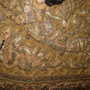 May include: Close-up of an ornate textile with intricate gold and bronze embroidery. The design features detailed figures and swirling patterns, with accents of purple and black. The edges are bordered with a beaded trim.