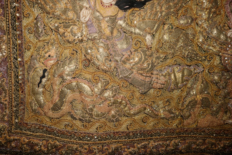 May include: Close-up of an ornate textile with intricate gold and bronze embroidery. The design features detailed figures and swirling patterns, with accents of purple and black. The edges are bordered with a beaded trim.