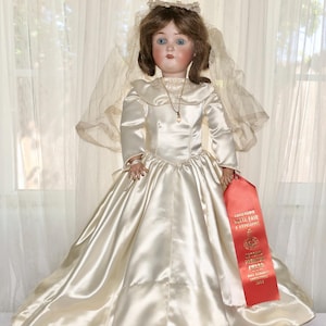 May include: A vintage porcelain doll wearing a satin wedding dress with a long skirt and a veil. The doll has blue eyes and brown hair. A red ribbon with gold text is held in the doll's hand.