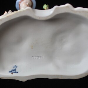 Puede incluir: La parte inferior de una figurita de porcelana con una marca azul y blanca que dice "Goebel" y el n&uacute;mero "1128/11".
