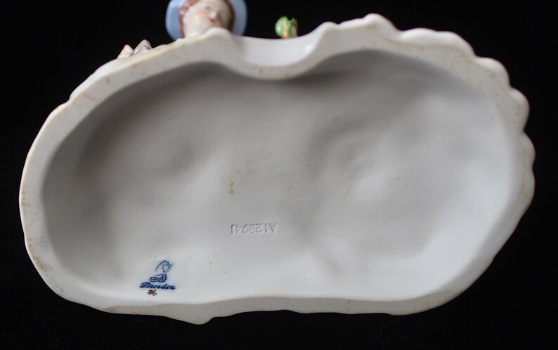 Puede incluir: La parte inferior de una figurita de porcelana con una marca azul y blanca que dice "Goebel" y el n&uacute;mero "1128/11".