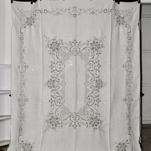 May include: A white linen curtain with a gray embroidered design featuring a floral pattern and geometric shapes. The curtain is hanging from a wooden rod.
