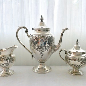 May include: A silver tea set with an ornate design. The set includes a teapot, creamer, and sugar bowl.