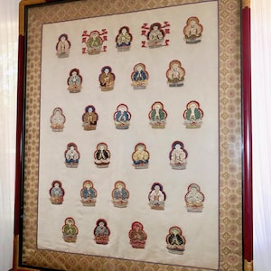 May include: Framed artwork featuring a series of embroidered figures in various colors. The figures are arranged in rows on a cream-colored background, framed with a red and gold border. The artwork is displayed on a wooden surface.