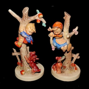 May include: Two ceramic figurines depicting children climbing trees, with dogs at the base. The figurines feature detailed painting with a neutral color palette, including red, blue, and brown. The figurines are on a round base.