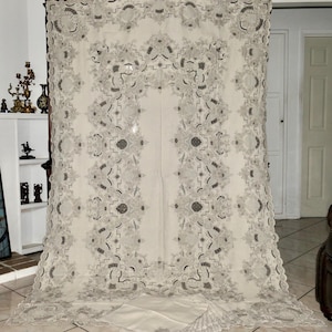 May include: A large, white linen tablecloth with intricate embroidered floral patterns in a silver thread. The tablecloth has a scalloped edge and is shown folded on a tiled floor.