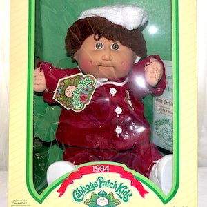 May include: A 1984 Cabbage Patch Kids doll with brown yarn hair, wearing a white knit hat and a maroon long-sleeved outfit with white buttons. The doll is inside a yellow and green cardboard box with the text Cabbage Patch Kids and I'm unique. I'm adoptable!