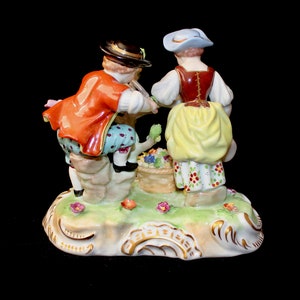 Puede incluir: Una figurita de porcelana de una pareja con ropa tradicional, el hombre con una chaqueta roja y la mujer con un vestido amarillo, de pie sobre una base verde con flores.