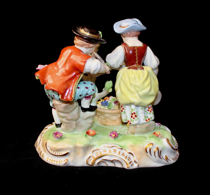 Puede incluir: Una figurita de porcelana de una pareja con ropa tradicional, el hombre con una chaqueta roja y la mujer con un vestido amarillo, de pie sobre una base verde con flores.