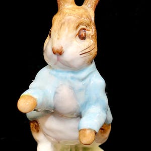 May include: A ceramic figurine of a brown rabbit standing on a small circular base. The rabbit has long ears, dark eyes, and is wearing a light blue jacket. The figure features painted details including a white chest and brown fur accents.