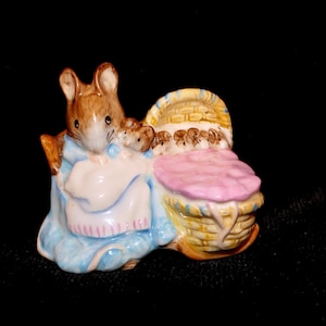 May include: A ceramic figurine depicting a brown mouse wearing a blue garment and a white apron, standing beside a yellow woven basket with a pink blanket inside. The mouse has a smaller mouse figure resting near its shoulder, all in a glossy finish.