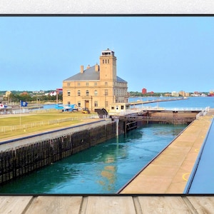 May include: A view of a lock on a canal with a large building in the background. The building is a light brown color with a tall tower. The canal is filled with water and there is a small boat in the distance.