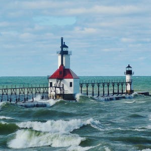 May include: Two white lighthouses with red roofs stand on a pier extending into a choppy, blue-green sea. The lighthouses have black and white striped towers and are connected by a wooden walkway.
