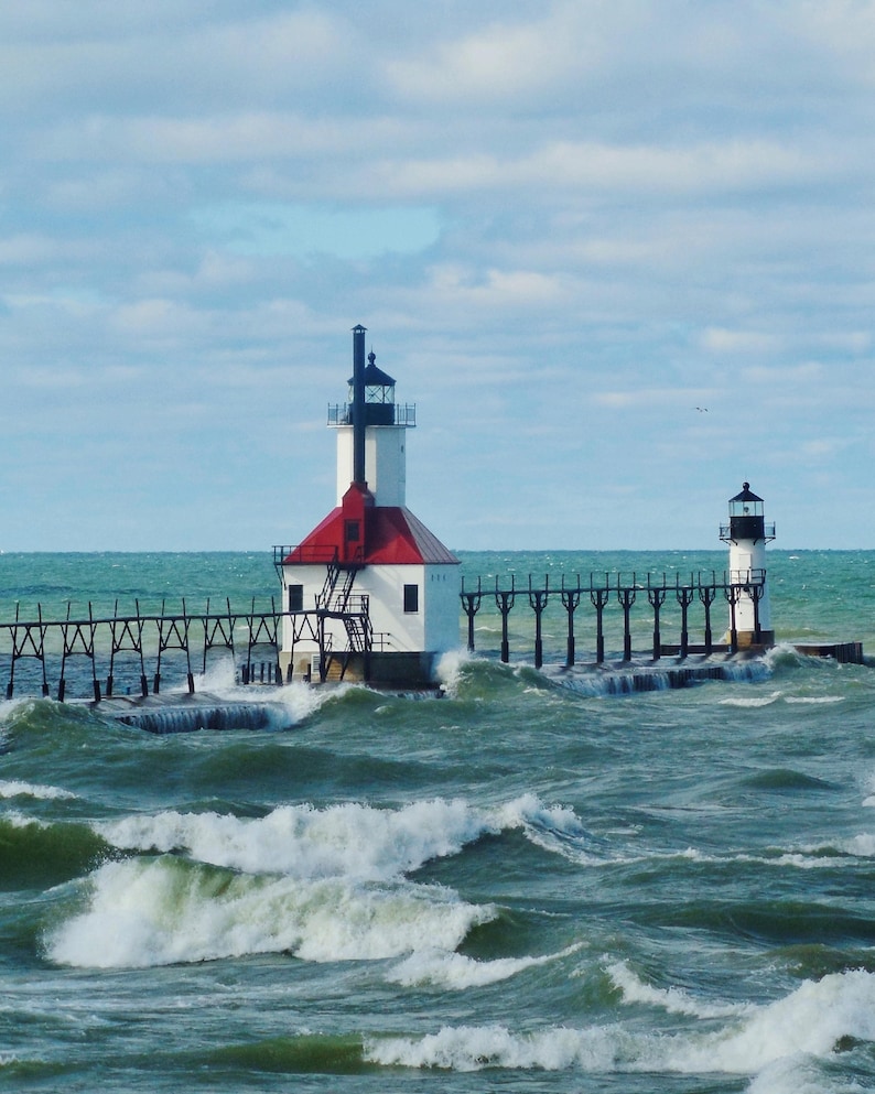 May include: Two white lighthouses with red roofs stand on a pier extending into a choppy, blue-green sea. The lighthouses have black and white striped towers and are connected by a wooden walkway.